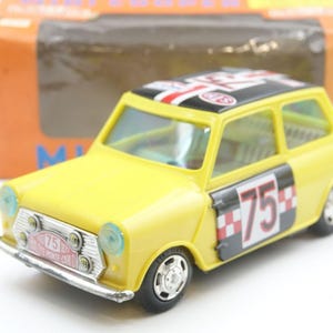 May include: A yellow toy car with a black roof featuring a Union Jack design. The car has the number 75 on the side and a silver front bumper. The car is in front of a brown and orange box.