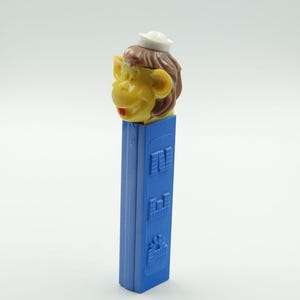 May include: A plastic Pez candy dispenser featuring a yellow monkey head with brown hair and a small white sailor hat. The dispenser body is a solid blue rectangular column with the word PEZ embossed vertically in a block font on the side.