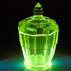 Könnte beinhalten: Ein leuchtend grünes Glasgefäß mit Deckel, vor schwarzem Hintergrund. Das Gefäß hat ein geometrisches Design mit facettiertem Körper und Deckel sowie einem spitzen Knauf. Das Glas scheint Uranglas zu sein und strahlt hellgrün.