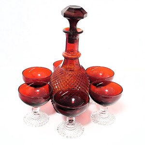 May include: A vintage ruby red glass decanter with a faceted stopper, surrounded by six matching stemmed glasses. The decanter and glasses have a diamond pattern, creating a classic glassware set.
