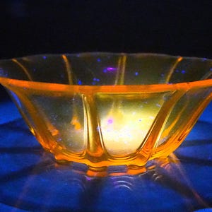 May include: A translucent orange glass bowl with a scalloped rim and vertical ribbed detailing. The bowl emits a bright glow under ultraviolet light, revealing a textured surface with small, scattered blue fluorescent specks.