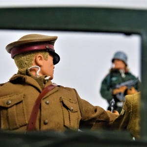 May include: A plastic action figure wearing a tan military uniform and a matching peaked cap with a dark brim. The figure is positioned in the foreground, facing away from the camera, while a second figure in a green uniform stands in the distance.