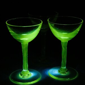 Puede incluir: Dos copas de cóctel vintage de color verde brillante con tallo y base redonda. Las copas están iluminadas sobre un fondo negro, resaltando su color y forma únicos. Una tercera copa es visible en el fondo.