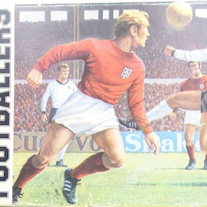 Puede incluir: Ilustración en caja de un kit de modelo de la serie deportiva, que representa un partido de fútbol. La caja muestra a dos jugadores con uniformes rojos y blancos pateando un balón. El texto "FOOTBALLERS" está impreso.