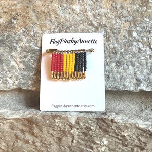 May include: A pin featuring a design of red, yellow, and black beads, resembling a flag. The pin is attached to a white card with the text "FlagPinsbyAnnette" and the Etsy shop URL. The pin is made with gold-colored safety pins.