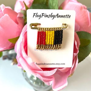 May include: A gold-colored safety pin with a beaded flag design. The flag features red, yellow, and black beads, representing the colors of the Belgian flag. The card reads "FlagPinsbyAnnette" and "flagpinsbyannette.etsy.com".