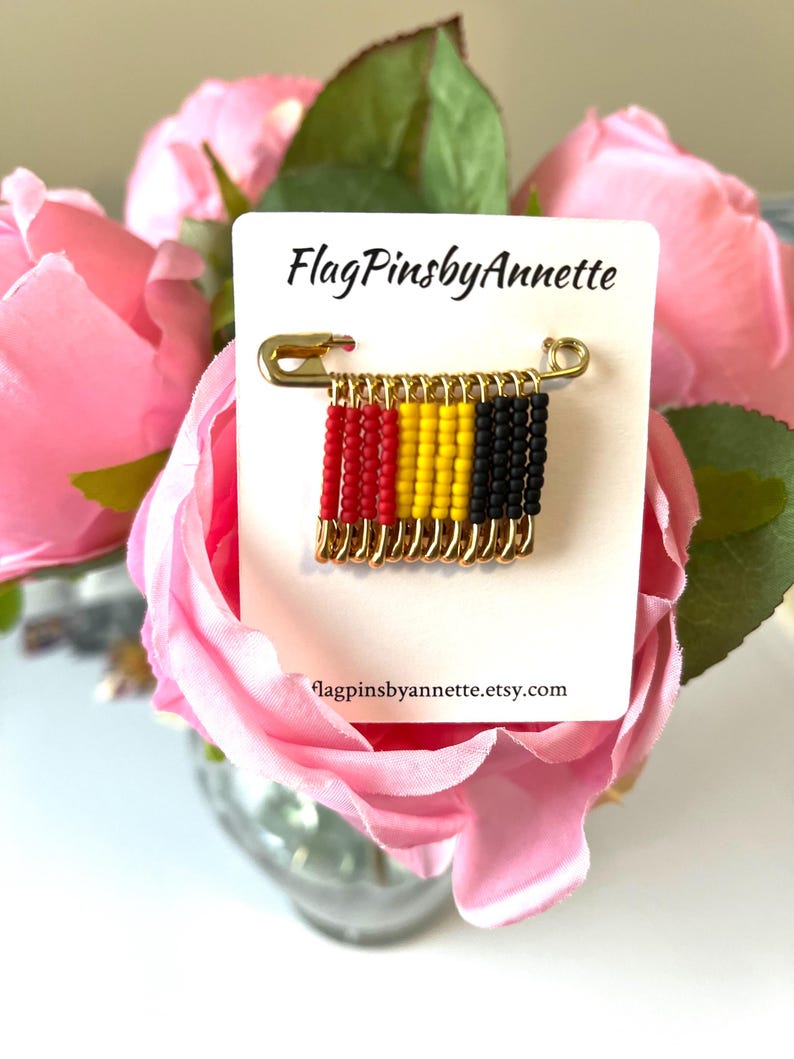 May include: A gold-colored safety pin with a beaded flag design. The flag features red, yellow, and black beads, representing the colors of the Belgian flag. The card reads "FlagPinsbyAnnette" and "flagpinsbyannette.etsy.com".