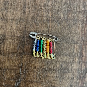 May include: A set of six rainbow-colored safety pins with gold-colored heads attached to a silver safety pin.
