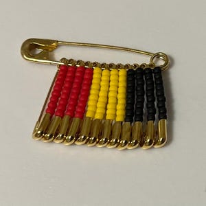 May include: A gold-toned safety pin with a beaded design. The beads are arranged in vertical rows of red, yellow, and black, creating a flag-like pattern. The pin is a decorative accessory.