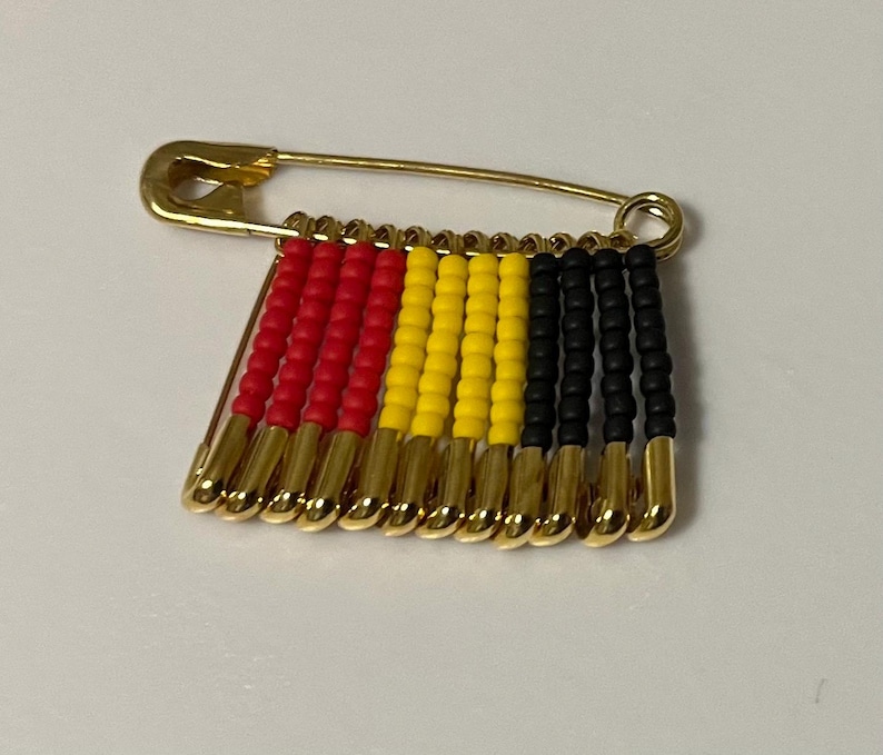 May include: A gold-toned safety pin with a beaded design. The beads are arranged in vertical rows of red, yellow, and black, creating a flag-like pattern. The pin is a decorative accessory.