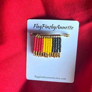 May include: A handmade pin featuring a miniature flag design made of red, yellow, and black beads. The pin is attached to a gold-colored safety pin and mounted on a white card with the text "FlagPinsbyAnnette" and a website address.