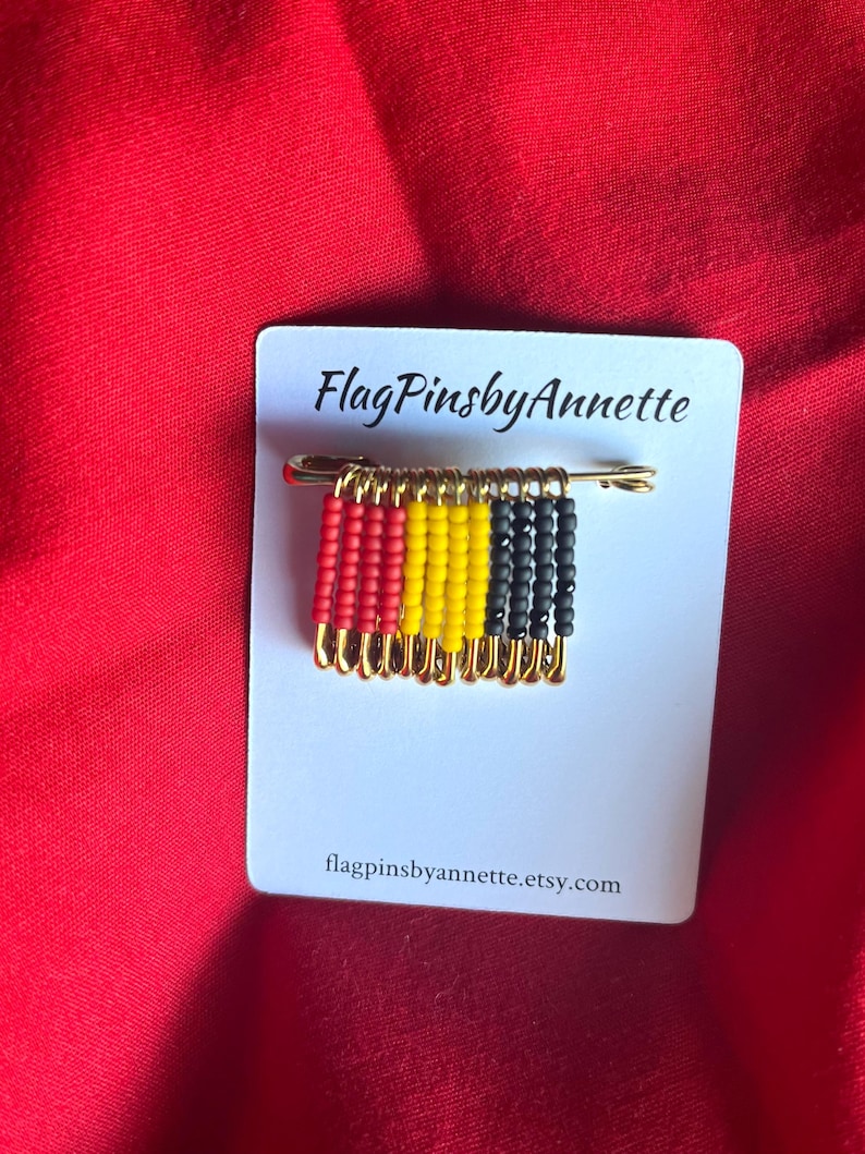 May include: A handmade pin featuring a miniature flag design made of red, yellow, and black beads. The pin is attached to a gold-colored safety pin and mounted on a white card with the text "FlagPinsbyAnnette" and a website address.