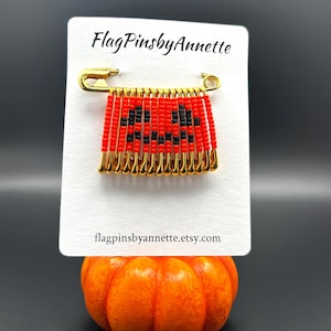 May include: A gold safety pin brooch featuring a row of smaller gold safety pins hanging from it. Each small pin is strung with red and black beads arranged to form a jack-o'-lantern face. The display card reads FlagPinsbyAnnette.