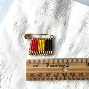 May include: A gold-toned safety pin with a beaded design in red, yellow, and black. The pin is approximately 1 inch long. The design resembles a flag. The pin is on a white, textured surface.
