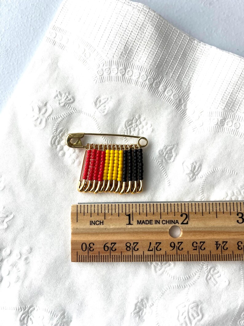 May include: A gold-toned safety pin with a beaded design in red, yellow, and black. The pin is approximately 1 inch long. The design resembles a flag. The pin is on a white, textured surface.