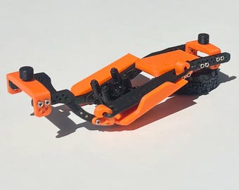 May include: A small, orange and black model vehicle chassis. The chassis has an orange frame with black detailing, including what appears to be a small tire. The model is designed to be a miniature version of a larger vehicle.