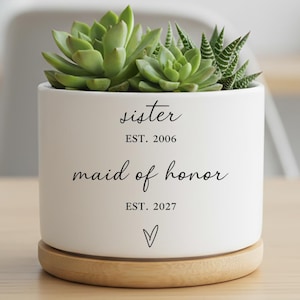 May include: A white ceramic planter with a succulent plant. The planter has the words "sister" and "maid of honour" printed on it, along with the years 2006 and 2027. The planter sits on a round, wooden base.