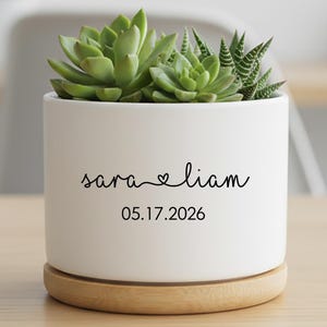 May include: A white ceramic planter with a wooden base, holding green succulents. The planter has the names "sara" and "liam" with a heart symbol, and the date "05.17.2026" in black script. A decorative item for home decor.