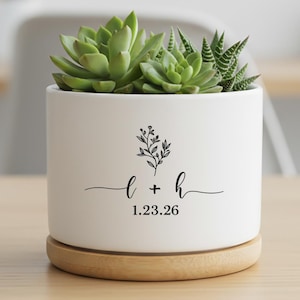 Może przedstawiać: Biała ceramiczna donica z drewnianą podstawą, zawierająca zielone sukulenty. Donica posiada czarny kwiatowy wzór oraz inicjały "L + h" z datą "1.23.26". Donica stoi na jasnobrązowej powierzchni.