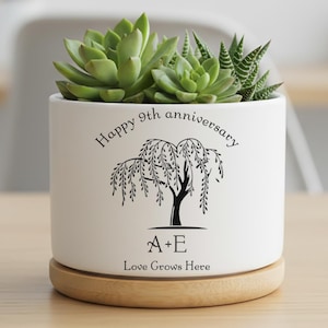 May include: White ceramic planter with a wooden base, holding green succulents. The planter is decorated with a black weeping willow tree design and the text "Happy 9th anniversary", "A+E", and "Love Grows Here".