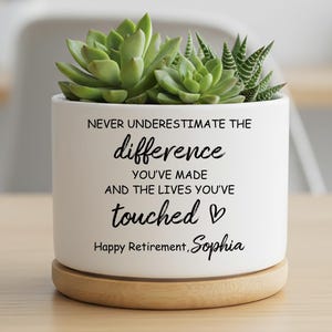 Peut inclure: Un pot de fleurs en céramique blanche avec une plante succulente. Le pot porte l'inscription "NEVER UNDERESTIMATE THE difference YOU'VE MADE AND THE LIVES YOU'VE touched" et "Happy Retirement, Sophia". Il repose sur une base en bois.