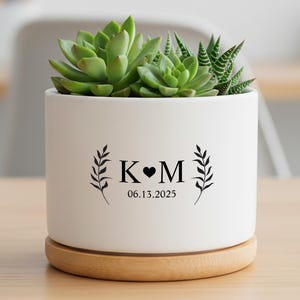 May include: A white ceramic planter with a succulent plant, resting on a wooden saucer. The planter features black text with initials and a date. The succulent has green leaves. The planter is on a light brown wooden table.