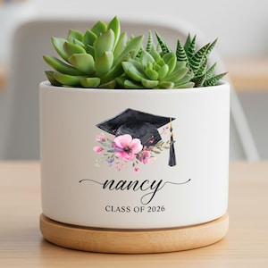 May include: A white ceramic planter with a graduation cap and floral design, holding green succulents. The planter has the name "nancy" and "CLASS OF 2026" printed on it. It sits on a round wooden base.