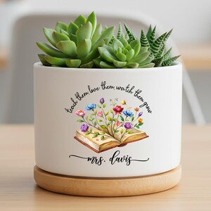 May include: White ceramic planter with a succulent plant. The planter has a wooden base and features a floral book design with the text "teach them love them watch them grow" and "mrs. davis".