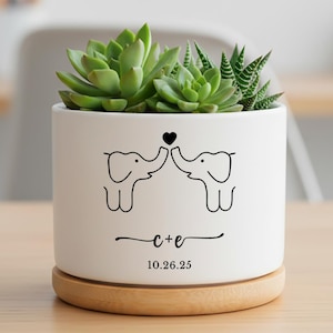Puede incluir: Maceta de cerámica blanca con base de madera, con una planta suculenta. La maceta tiene un diseño de elefantes negros con un corazón, iniciales y una fecha. La maceta mide aproximadamente 10 cm de alto.