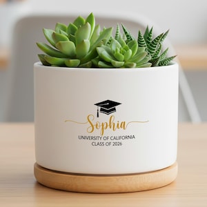 May include: A white ceramic planter with a succulent plant, resting on a wooden saucer. The planter features a graduation cap graphic and the name "Sophia" with "University of California Class of 2026" in gold lettering. The succulent has green leaves.