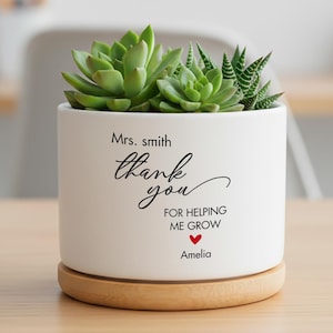 May include: A white cylindrical ceramic planter with a light wood base, containing green succulents. The front features black text reading Mrs. Smith, thank you for helping me grow, with a small red heart above the name Amelia printed below.
