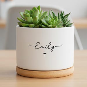 Puede incluir: Una maceta cilíndrica de cerámica blanca con base de madera clara, que contiene plantas suculentas verdes. El frente de la maceta presenta el nombre Emily en letra cursiva negra, situado sobre un pequeño símbolo de cruz negra impreso.