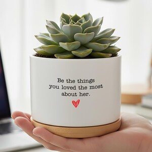 May include: A white ceramic planter with a succulent plant, sitting on a wooden base. The planter has the text "Be the things you loved the most about her." with a red heart. The plant is green.
