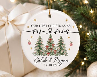 以下が含まれることがあります： 「OUR FIRST CHRISTMAS AS MR & MRS」という文字とハートのシンボルが描かれた白い円形のセラミック製オーナメント。その下には赤いリボンが付いた3本の緑の松の木が描かれ、続いて「Caleb & Megan」という名前と「18.12.26」という日付が記されています。