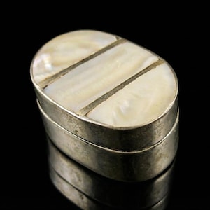 May include: An oval silver box with a mother-of-pearl lid. The lid is divided into three sections by thin silver lines. The box is reflected in the black surface below. A small decorative container.