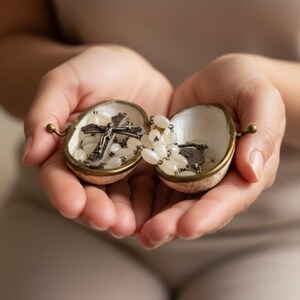 Open vintage mother-of-pearl clam shell box with brass trim, held in two hands and filled with a small white-beaded Catholic rosary and crucifix