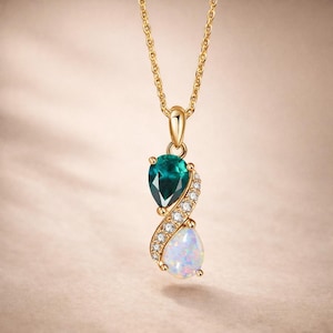 Puede incluir: Collar dorado con colgante. El colgante tiene una gema verde esmeralda en forma de lágrima sobre un ópalo blanco, también en forma de lágrima. Pequeñas piedras transparentes añaden brillo. El collar cuelga de una fina cadena dorada.