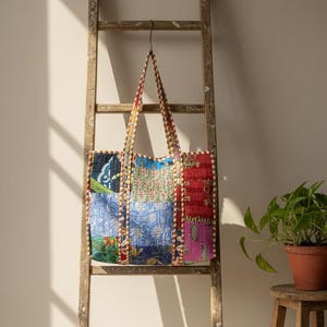 May include: A patchwork tote bag with a variety of colorful fabric panels, including blue, red, and floral patterns. The bag hangs from a wooden ladder, with a small plant on a wooden stool in the background. The bag has a matching patterned handle.