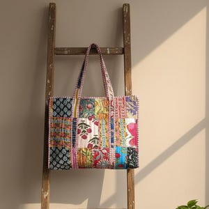 Könnte beinhalten: Eine Patchwork-Tragetasche mit einer Vielzahl farbenfroher Blumen- und geometrischer Muster. Die Tasche hat stabile Griffe und wird auf einer Holzleiter präsentiert. Das Design umfasst rote, blaue, grüne und gelbe Farbtöne.