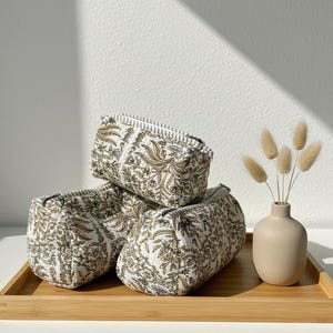 May include: Three small, rectangular cosmetic bags with a white and gold floral pattern are stacked on a wooden tray. The bags have a zipper closure and a white trim. A small beige vase with dried flowers sits on the tray.