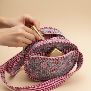 May include: A floral-patterned quilted bag with a zip closure and a long strap. The bag has a design of pink flowers and green leaves on a grey background. A gold lipstick tube is being placed inside the bag.