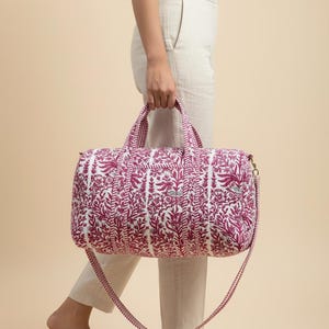 May include: A white and magenta floral print duffel bag with a matching shoulder strap and handles. The bag is quilted and has a gold-colored zipper. The bag is being carried by a person wearing light-colored pants and sandals.