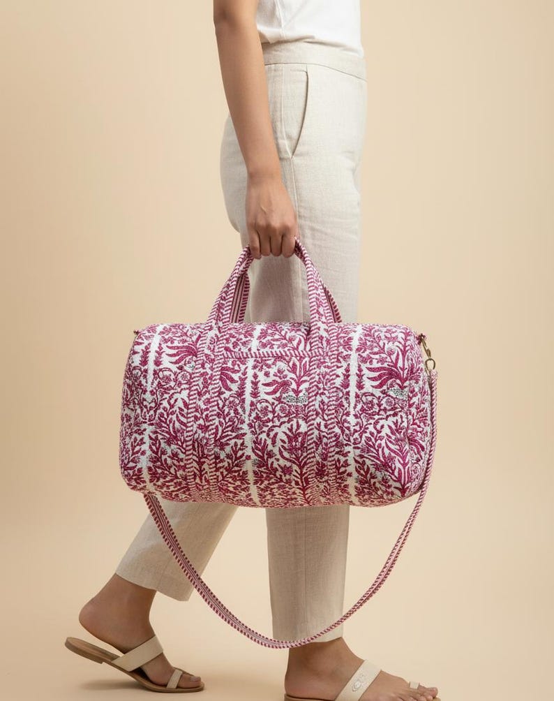 May include: A white and magenta floral print duffel bag with a matching shoulder strap and handles. The bag is quilted and has a gold-colored zipper. The bag is being carried by a person wearing light-colored pants and sandals.