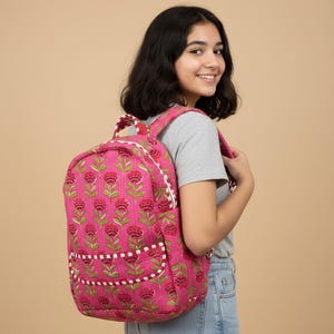 May include: A pink floral backpack featuring a pattern of red flowers and green leaves. The bag has white trim, a front pocket, and padded shoulder straps. The backpack is made of quilted fabric.