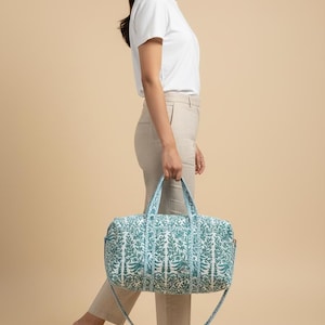 May include: A rectangular duffel bag with a teal and white floral pattern. The bag has two handles and a detachable shoulder strap. The bag is being carried by a person wearing a white t-shirt and tan pants.