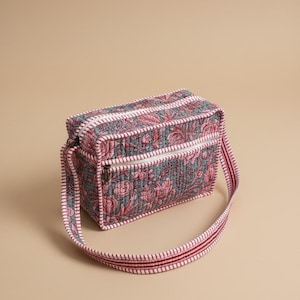 May include: A quilted crossbody bag with a floral pattern in shades of pink and gray. The bag has a rectangular shape with a zipper closure and a striped strap. The bag's dimensions are approximately 10 inches long, 6 inches high, and 4 inches wide.