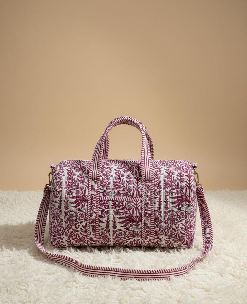 May include: A quilted travel bag with a floral pattern in shades of burgundy and white. The bag has a rectangular shape with a top handle and a detachable shoulder strap. The bag measures approximately 10 inches in length, 6 inches in height, and 6 inches in width.
