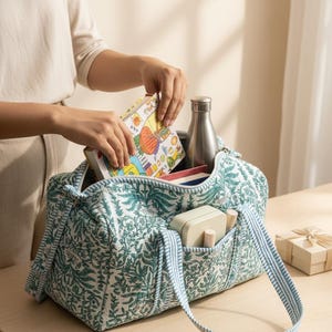 May include: A quilted tote bag with a green and white floral pattern, blue striped trim, and handles. The bag contains a book, a stainless steel water bottle, and other items. A small gift box sits nearby.