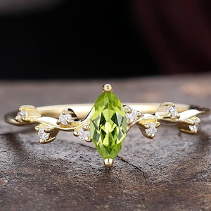 May include: A gold ring featuring a central marquise-cut peridot gemstone. The band is designed with a vine-like motif, accented by several small, round, clear white stones set along the branches extending from the center stone.