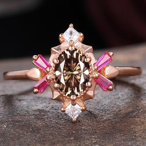 May include: Rose gold ring featuring a central oval-cut brown gemstone, surrounded by pink and white accent stones. The ring has a detailed, ornate setting with a vintage-inspired design. The band is a delicate rose gold color.